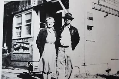 Two men in front of a building - one in suit and hat, one in overalls and cap