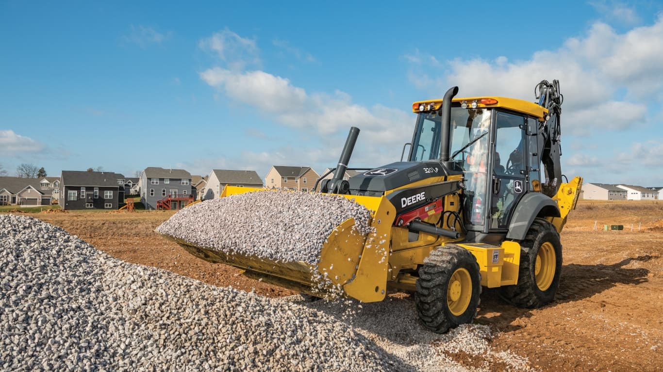 A 320P Backhoe hauling small rocks in the bucket with houses in the background. | 4re Equipment | ID: 320PFT4
