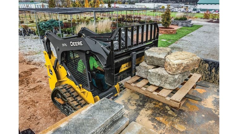 A 317G Compact Track Loader with pallet forks attachment lifting a pallet of large rocks at a nursery. | 4re Equipment