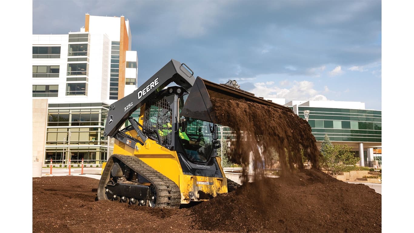 A 325G Compact Track Loader moving dirt at a jobsite with a commercial building in the background. | 4re Equipment | ID: 325gxt