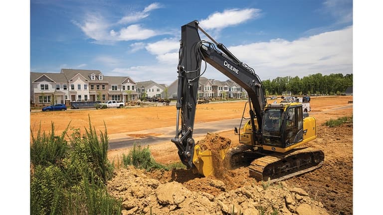 A 160P-Tier Excavator moving dirt at a worksite with a housing development in the background. | 4re Equipment