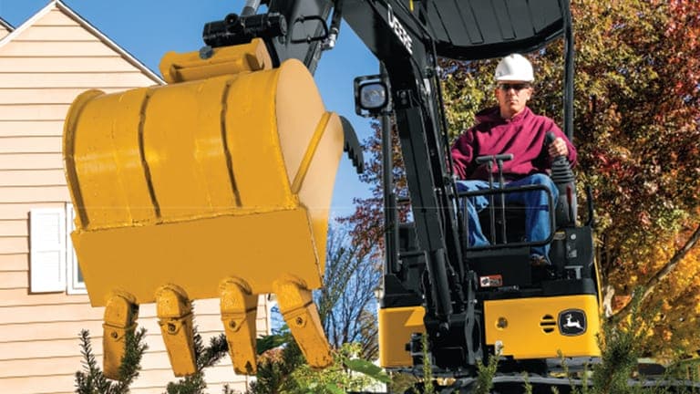 A close up of the 17P-Tier Excavator bucket hovering above plants with an operator and house in the background. | 4re Equipment