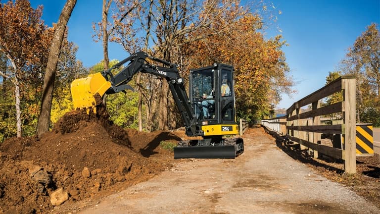 Man using 26G Excavator to stockpile dirt on a sidewalk with a wooden fence. | 4re Equipment