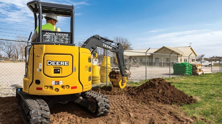 An operator moving topsoil to the right of a John Deere 30G Excavator in front of a chain link fence. | 4re Equipment