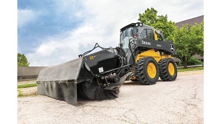 A 320G Skid Steer with broom attachment sweeps grass and dirt off a road. | 4re Equipment