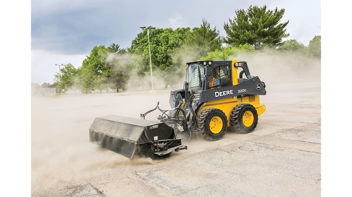 A 320G Skid Steer with pick-up broom attachment sweeps dirt and dust off a parking lot. | 4re Equipment | ID: 320gxt