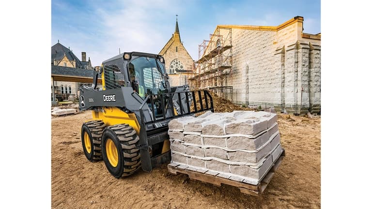 A 320G Skid Steer with pallet attachment transporting a pallet of stones with a church in the background. | 4re Equipment