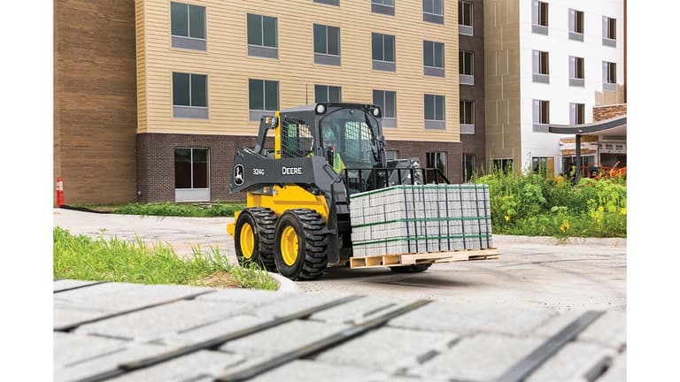 A 324G Skid Steer with pallet attachment transporting bricks in front of a building worksite. | 4re Equipment