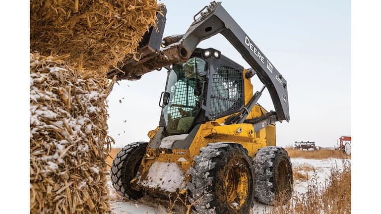 A 330G Skid Steer with bale spears attachment lifting a bale of snowy hay in a snowy field. | 4re Equipment